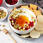 Honey Whipped Feta Dip with Pistachio and Pomegranate in a bowl next to crackers, a honey drizzler, pomegranate fruit, pomegranate seeds, chopped pistachios, and a kitchen towel.