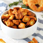 Rosemary Croissant Croutons in a bowl next to fresh rosemary, croissants, and scattered croutons.