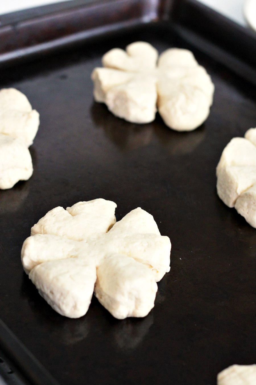 Uncooked biscuit dough cut into shamrock shapes spread out on a dark baking sheet.