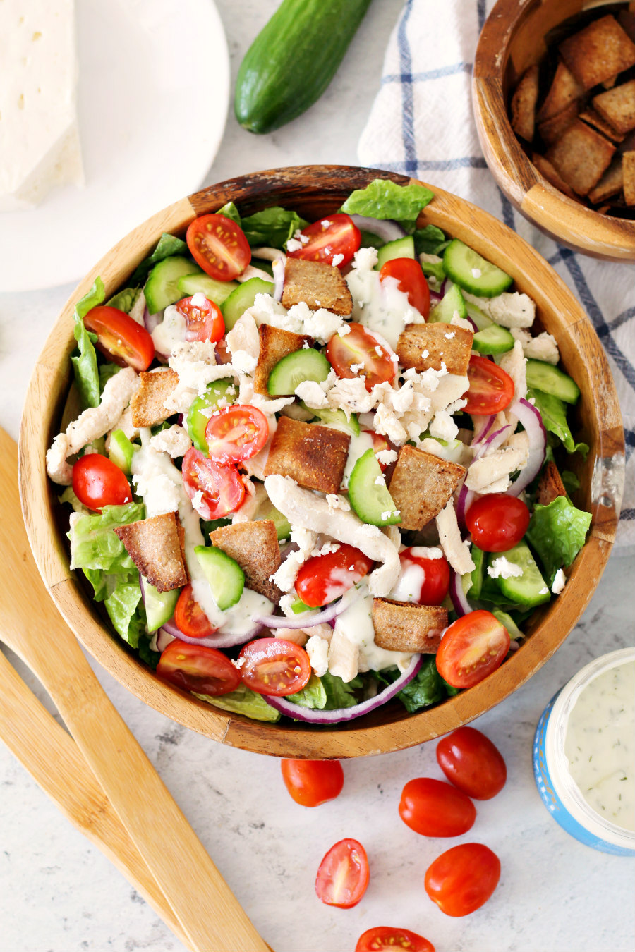 Chicken Gyro Salad with Pita Croutons in a wooden salad bowl. Wooden utensils, feta cheese, cucumber, pita croutons, tzatziki sauce, and cherry tomatoes in the background.