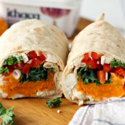 Sweet Potato and Veggie Hummus Wrap on a wooden board with a kitchen towel. Kale, sweet potato, feta cheese block, and hummus in the background.