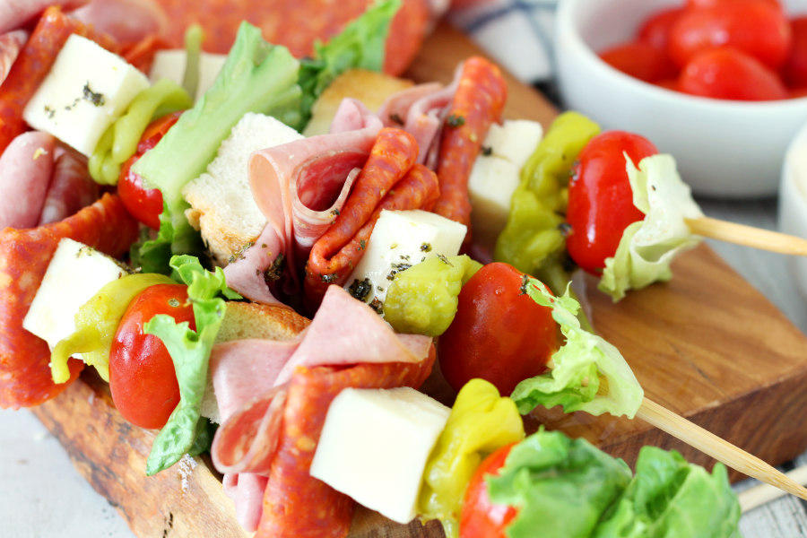 Italian Hoagie Skewers on a wooden board. Bowl of grape tomatoes in the background.