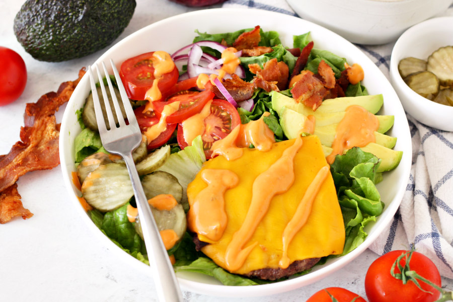Loaded Bacon Burger Bowl with fork on top. Bacon, tomato, avocado, and pickles arranged around it.