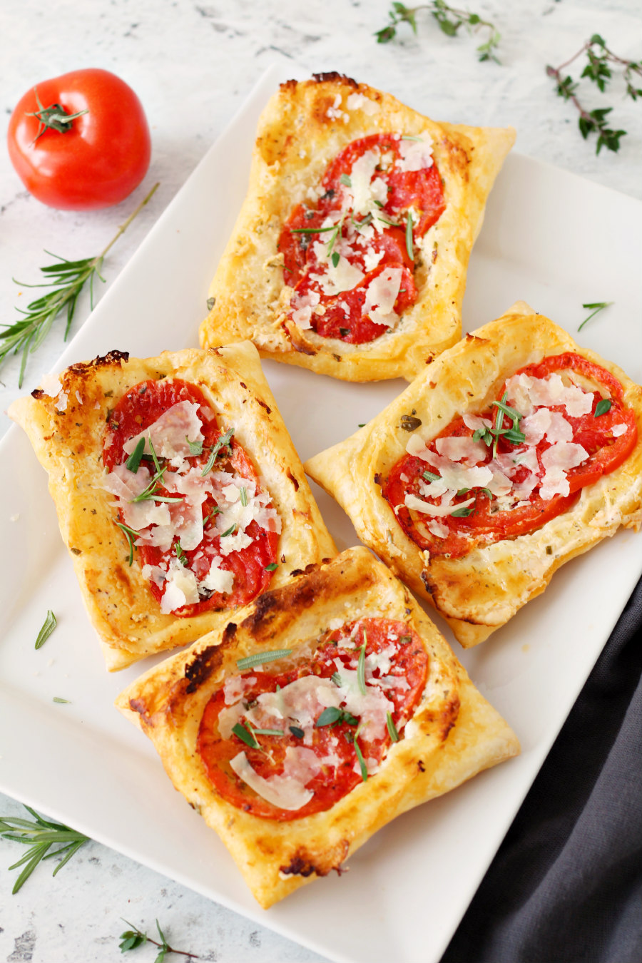 White plate of Upside Down Tomato and Herb Puff Pastry. Fresh herbs, tomato, and a kitchen towel surround the plate.