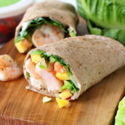Horizontal photo of Island Mango and Shrimp Wrap on a wooden board with ingredients in the background.