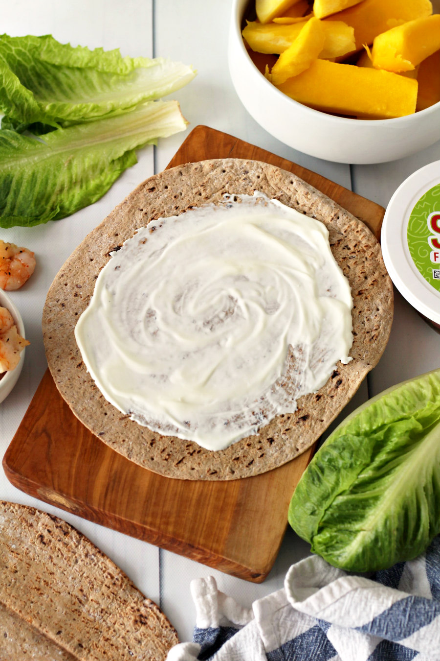Flat lay photo of soft tortilla with sour cream on a wooden board. Other Island Mango and Shrimp Wrap ingredients around it.