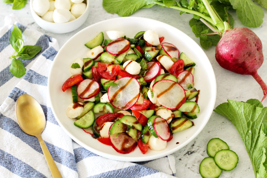 Horizontal photo of Tomato, Cucumber, and Radish Salad in a white serving dish. Spoon, cucumber slices, radish, mozzarella balls, and fresh basil surround the dish.