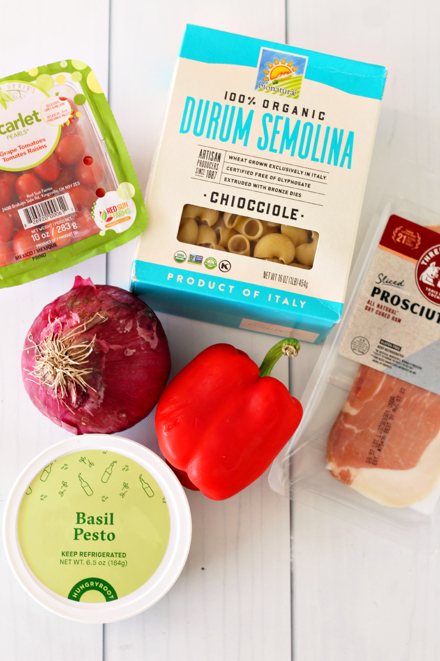 Flat lay photo of grape tomatoes, pasta, prosciutto, red onion, red bell pepper, and basil pesto.