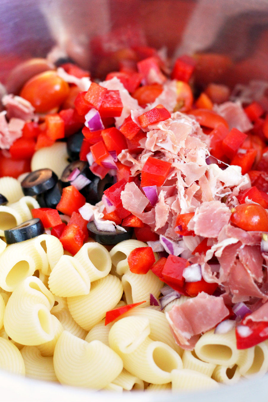 Chiocciole pasta in a mixing bowl with red bell pepper, onion, olives, grape tomatoes, and prosciutto.
