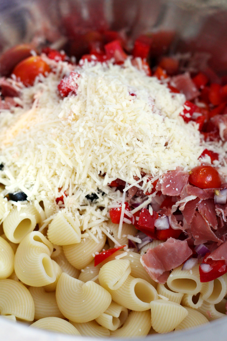 Chiocciole pasta in a mixing bowl with red bell pepper, onion, olives, grape tomatoes, and prosciutto with shredded Parmesan cheese on top.
