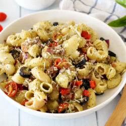 Horizontal photo of Pesto Pasta Salad with Prosciutto in a serving bowl. Fresh basil, grape tomatoes, a kitchen towel, and wooden spoon sit around the bowl.