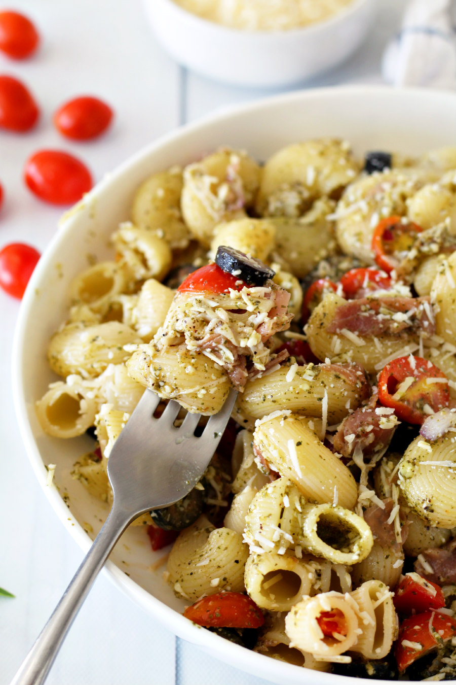 Forkful of Pesto Pasta Salad with Prosciutto sitting in a serving plate. Grape tomatoes and bowl of shredded Parmesan in the background.