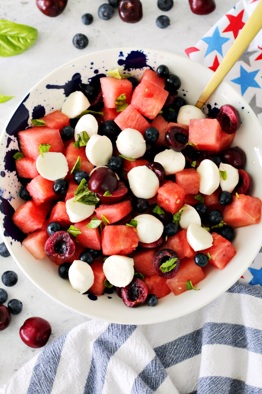 Flat lay photo of Watermelon Fruit Salad with Basil and Mozzarella in a serving bowl with a spoon. Fruit, basil, and patriotic towels sit around it.