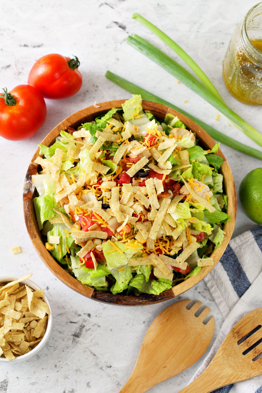 Flat lay photo of Southwestern Chopped Salad in a wooden salad bowl. Tomatoes, scallions, mason jar of dressing, lime, wooden utensils, and tortilla strips sit around the bowl.