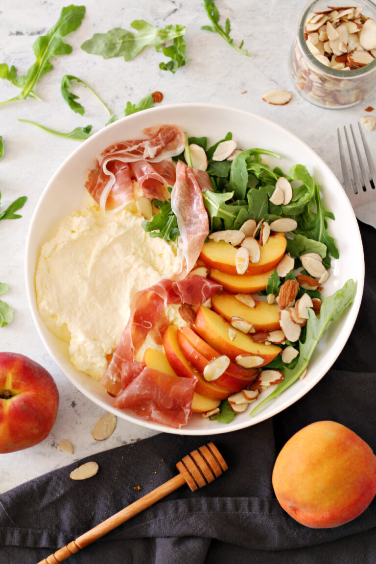Flat lay photo of Peach and Arugula Ricotta Bowl. Honey drizzler, peaches, arugula, and almonds sit around the bowl.