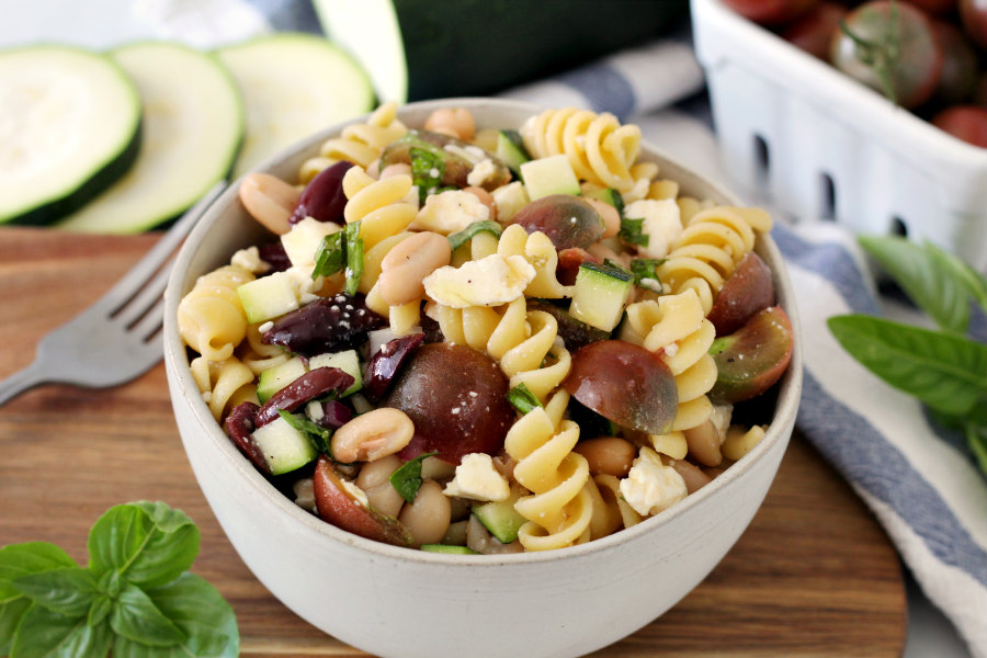 Horizontal photo of Summer Zucchini Pasta Salad sitting on wooden board with fresh basil, fork, zucchini, and cherry tomatoes in photo.