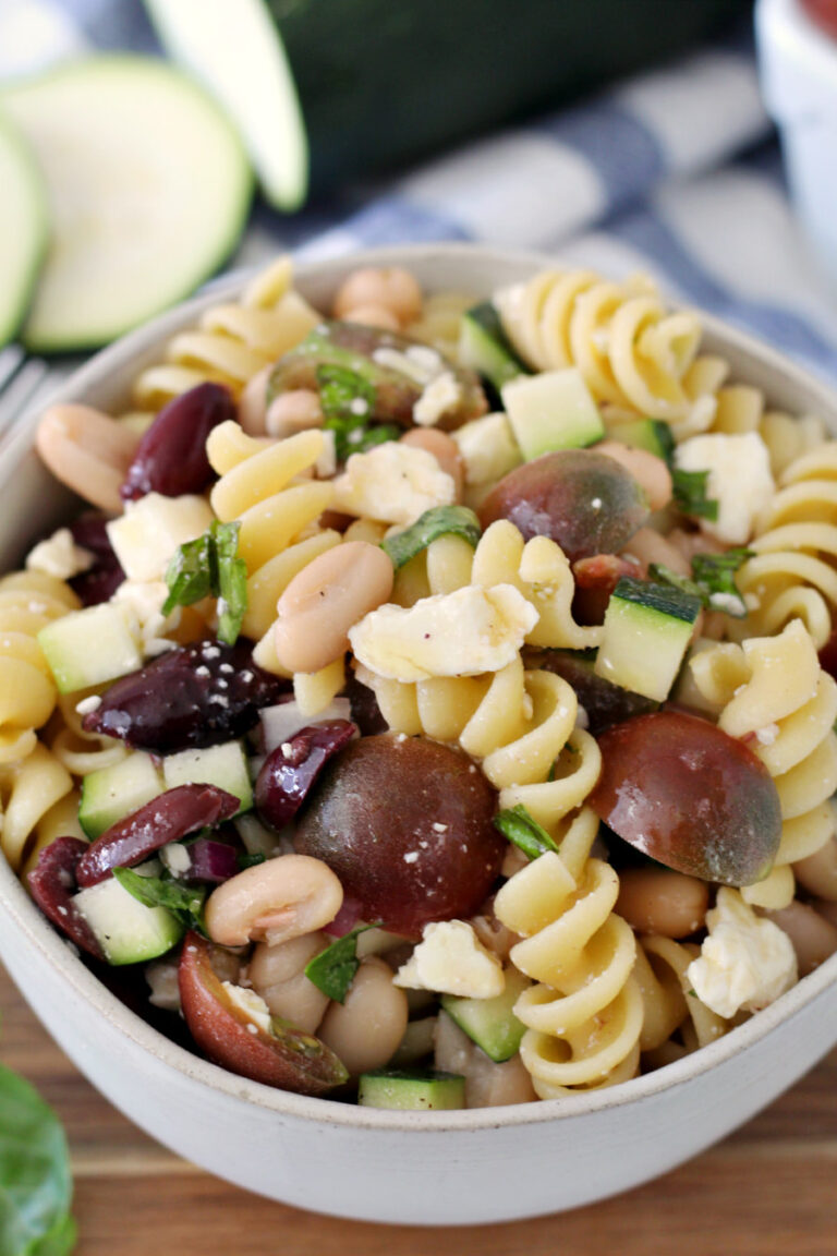 Summer Zucchini Pasta Salad in a bowl on a wooden board with fresh zucchini in the background.