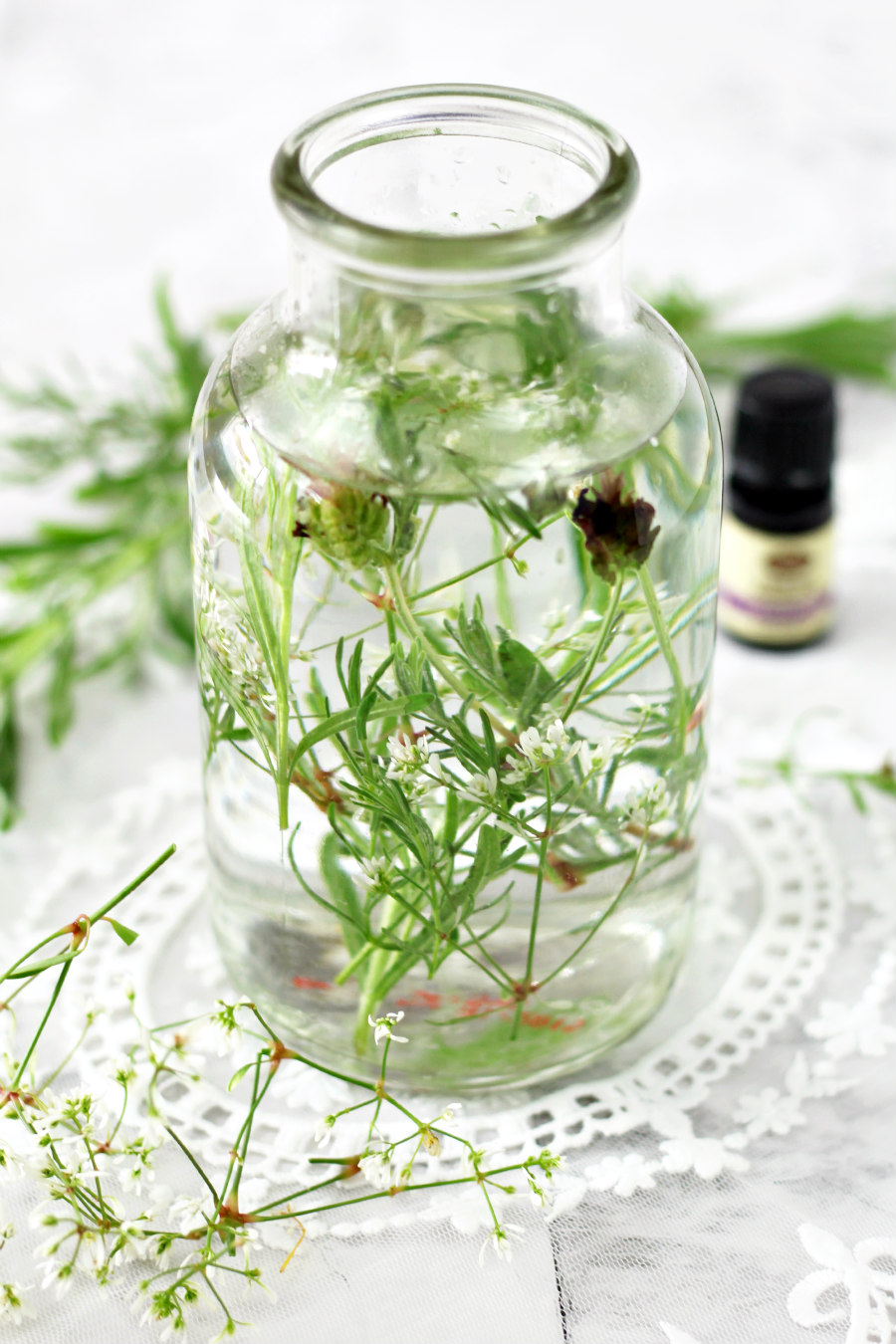 Botanical Herb Fragrance Diffuser in center of photo with fresh lavender sprigs and tiny white flower stems around it. A bottle of lavender essential oil sits in the background.