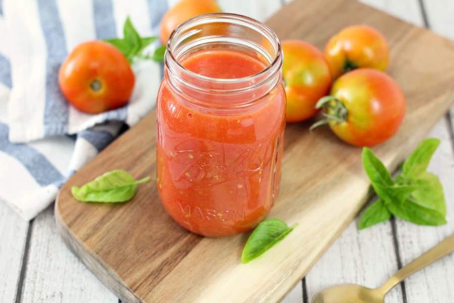 Horizontal photo of homemade garden crushed tomatoes in a mason jar. Jar sits on wooden board with basil and fresh tomatoes. Kitchen towel and spoon also in photo.