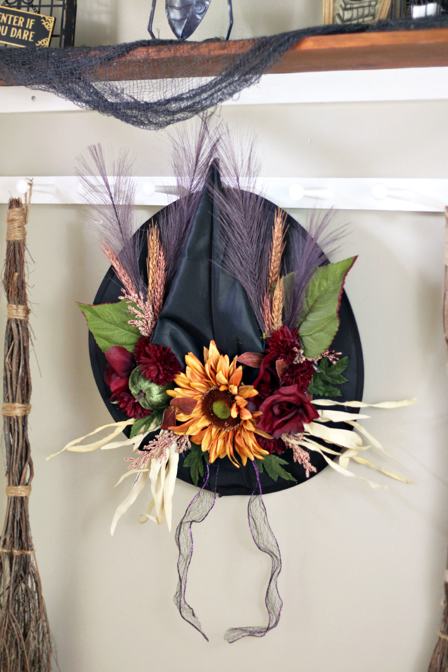 Fall Floral Witch Hat hung on hooks next to cinnamon broom and shelf of Halloween decorations.