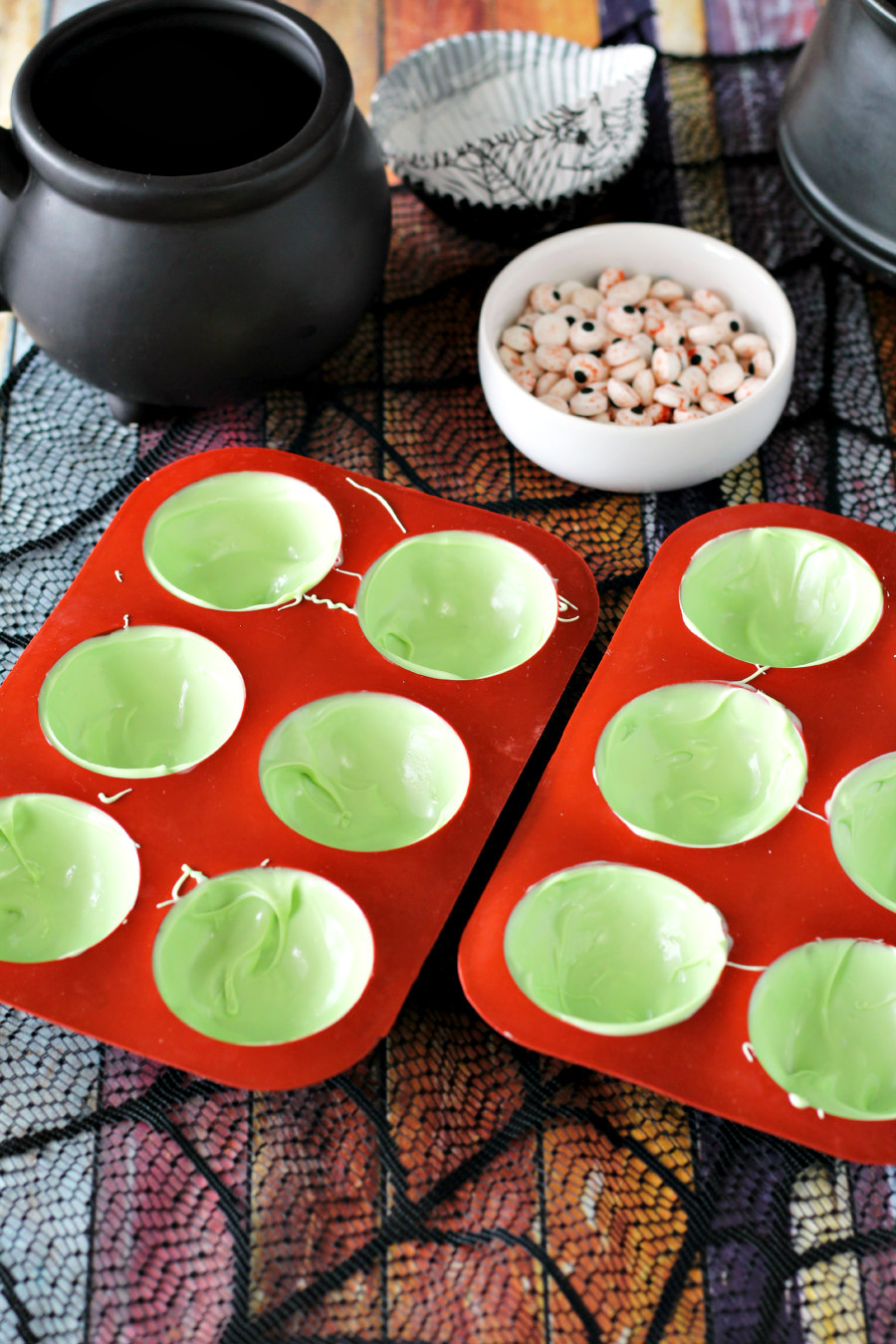 Light green candy melts spooned into silicone hot cocoa bomb molds. Cauldron mug, cupcake liners, and eyeball sprinkles in background.