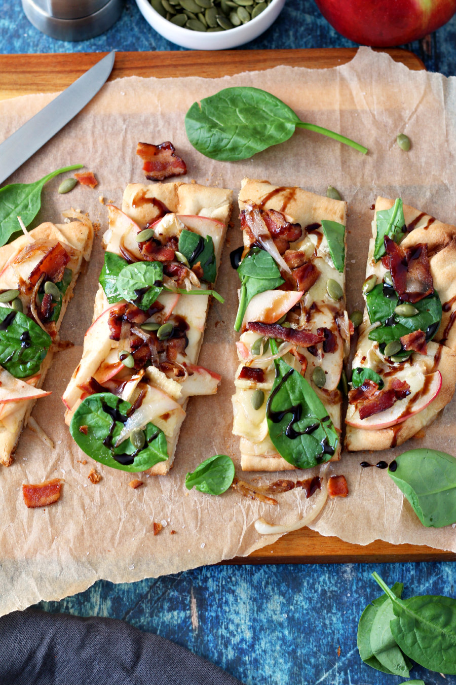 Flat lay photo of Apple, Bacon, and Brie Flatbread cut into pieces on a parchment lined wooden board. Knife, sits next to flatbread along with scattered baby spinach, crumbled bacon, and pepitas