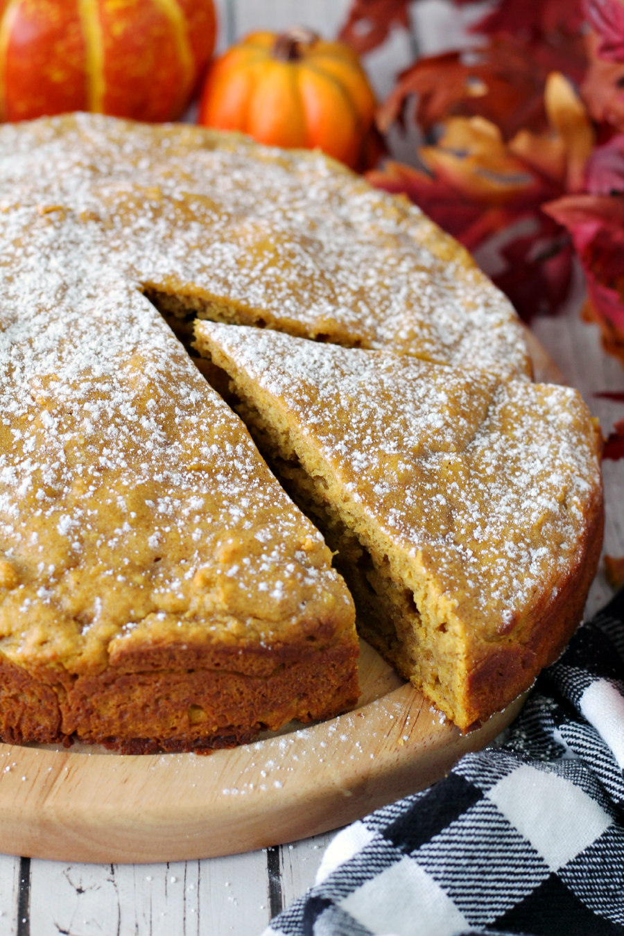 Close up of Pumpkin Spice Ricotta Cake with slice cut out. Checkered towel, pumpkins, and autumn leaves also in photo.