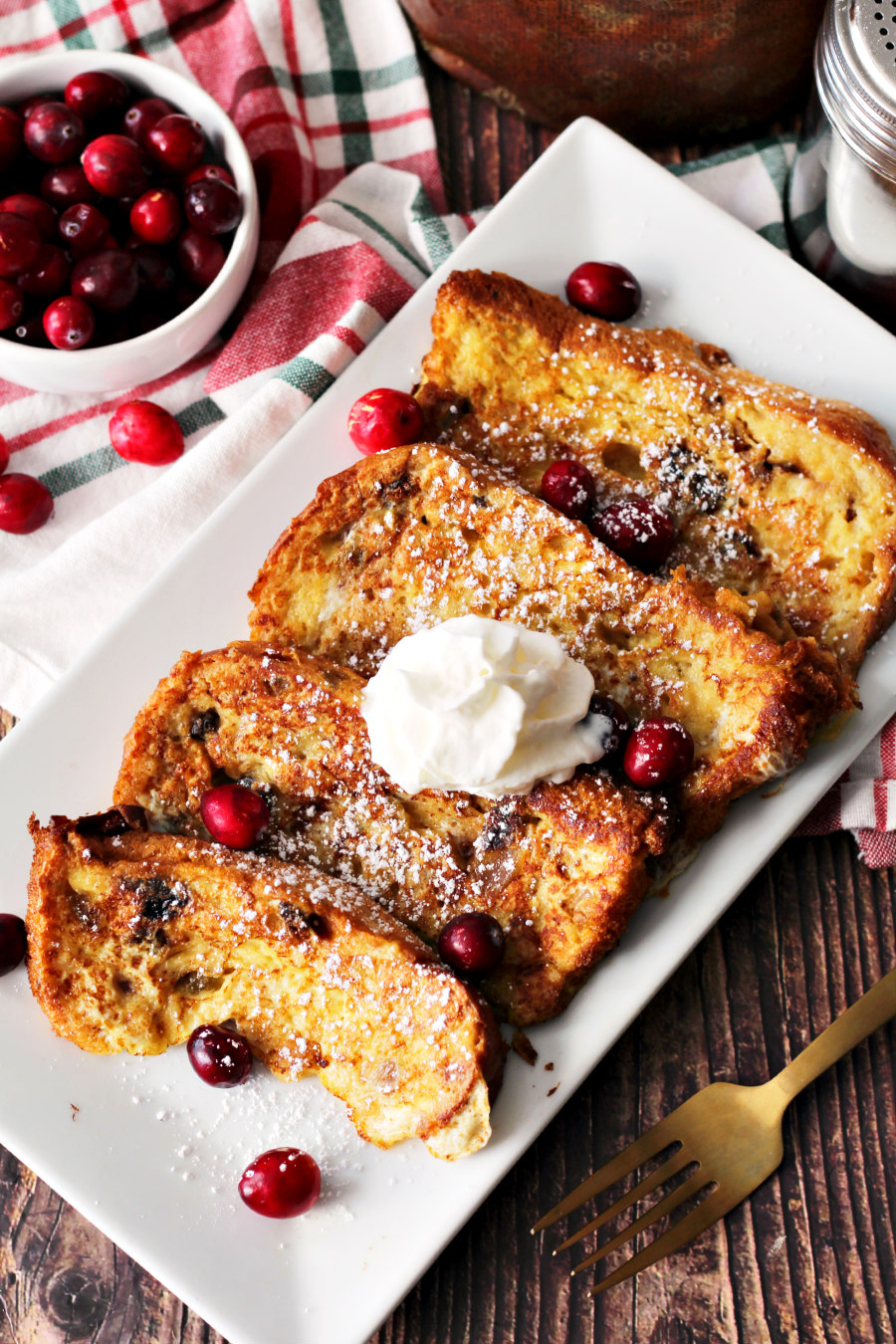 Flat lay photo of Panettone French Toast on white serving dish. Gold fork and small bowl of fresh cranberries also in photo.