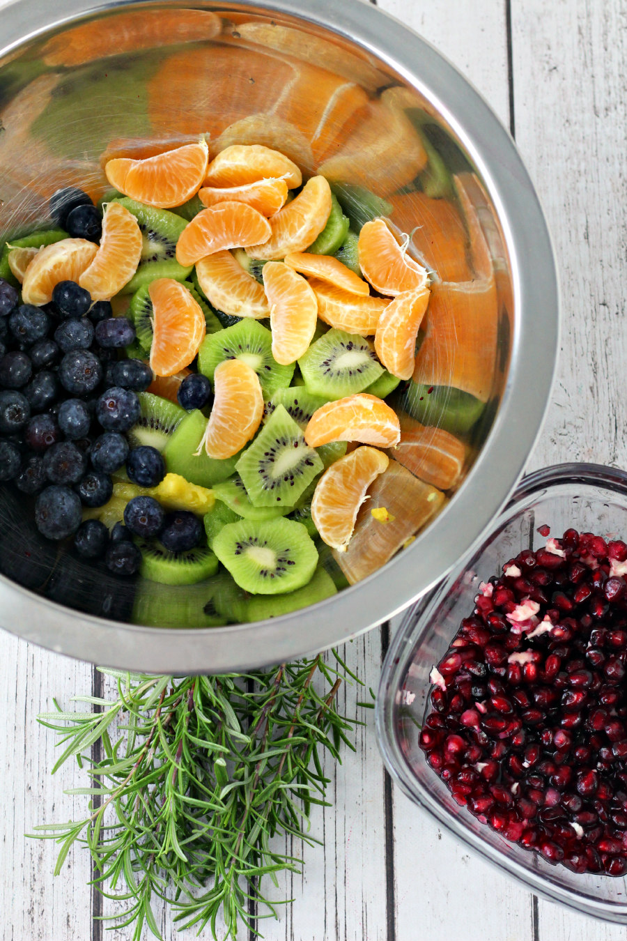 Prepared pineapple, clementines, blueberries, kiwis, and pomegranate arils in bowls. Sprigs of fresh rosemary sit alongside bowls.