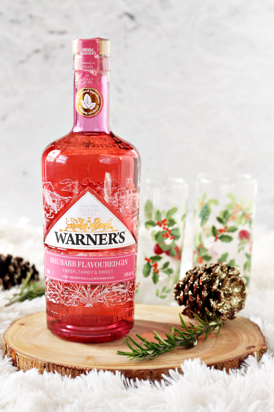 Bottle of Warner's Rhubarb Gin on wood slice with holly berry glasses in background. Fresh rosemary and sparkly pine cones also sit in photo.