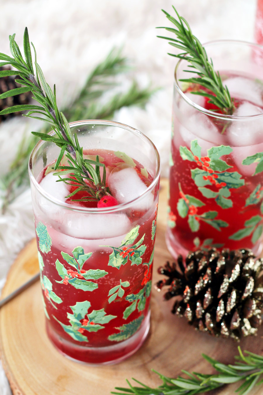 Rhubarb and Cranberry Gin and Tonic in holly berry glasses garnished with fresh rosemary sprigs. Glasses sit on wood slice with fresh rosemary and sparkly pine cones.