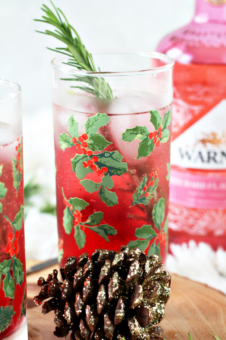 Rhubarb and Cranberry Gin and Tonic in holly berry glass garnished with fresh rosemary sprig. Bottle of Warner's Rhubarb Gin sits in background. Sparkly pine cone sits in front of glass.