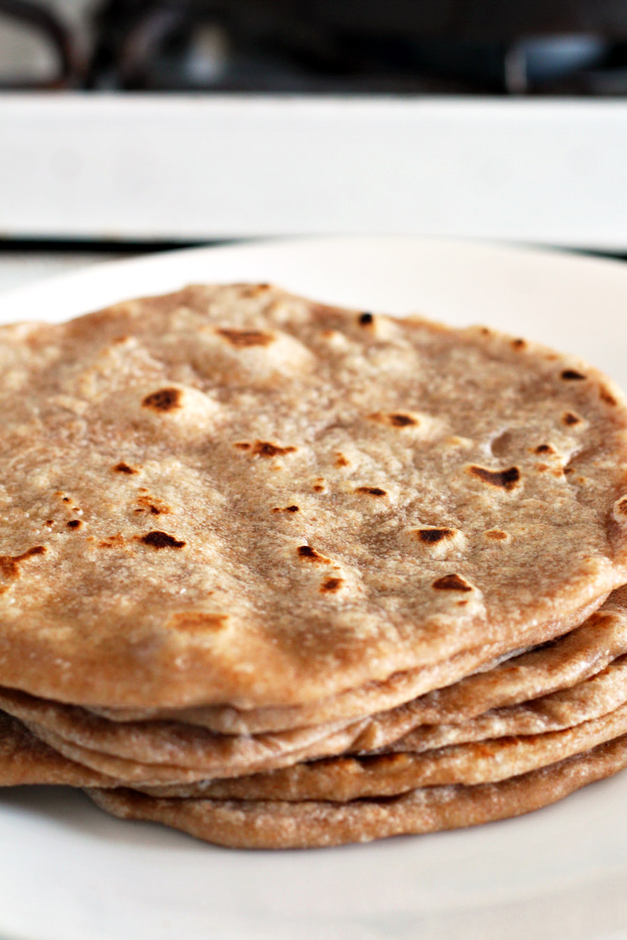 Half Whole Wheat Homemade Tortillas stacked on a white plate. Plate sits in front of gas stove.