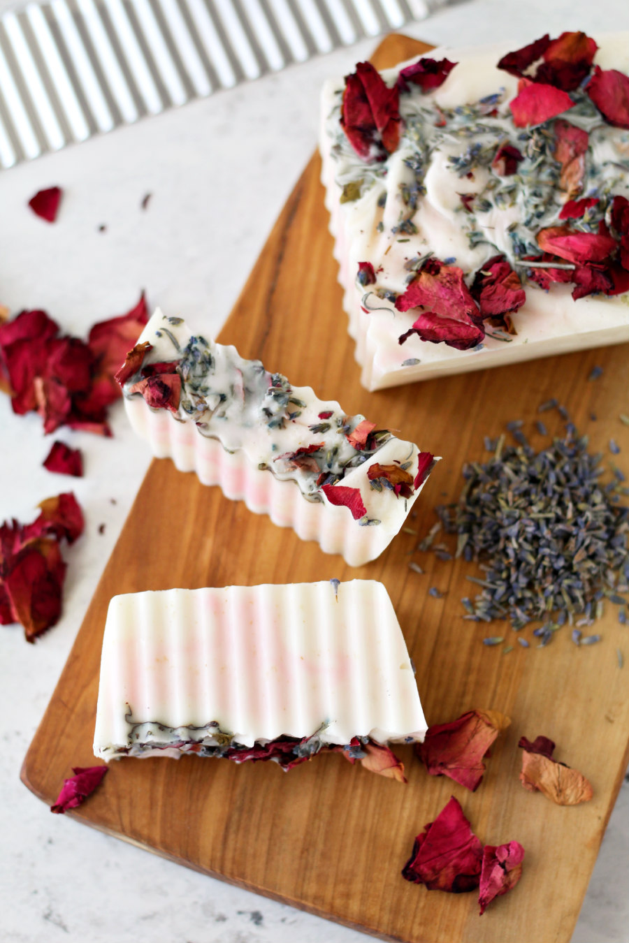 Rose Petal and Lavender Soothing Calming Floral Oatmeal Soap Bar from Holoka Home on Etsy. Soap loaf sits on wood board with soap bars, dried lavender, and dried rose petals. Soap cutter sits in background.