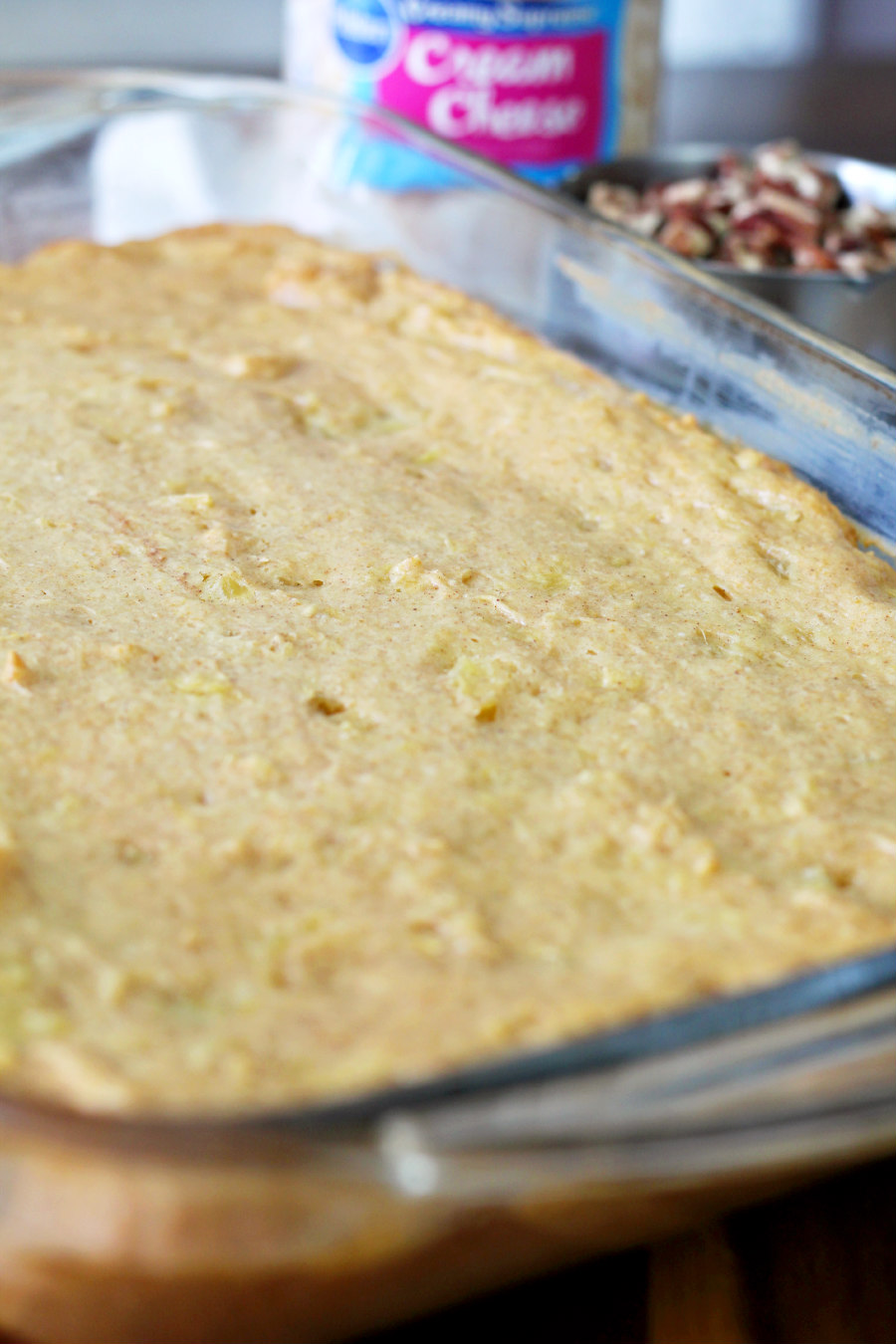 Hummingbird Pudding Dump Cake in glass baking pan. Container of cream cheese frosting and measuring cup of chopped pecans sit in background.