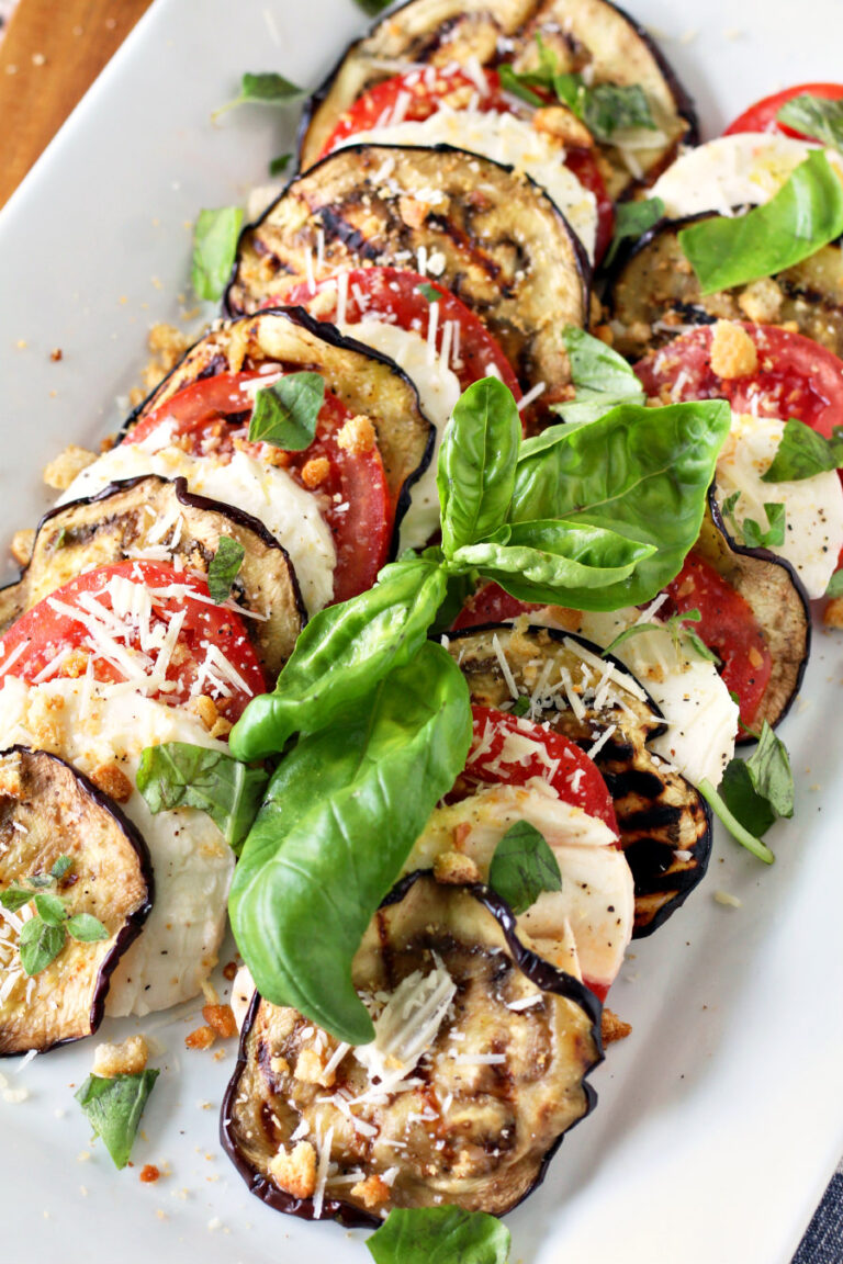 Layered grilled eggplant slices, sliced tomato, and fresh mozzarella sit on white serving platter. Salad is topped with fresh herbs, crushed croutons, shredded Parmesan, and a drizzle of balsamic glaze.