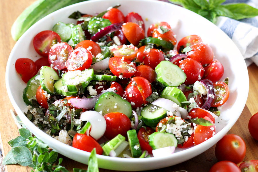 Horizontal photo of Fresh Tomato, Cucumber, and Herb Salad surrounded by blue and white striped kitchen towel, fresh herbs, mini cucumber, and cherry tomatoes.