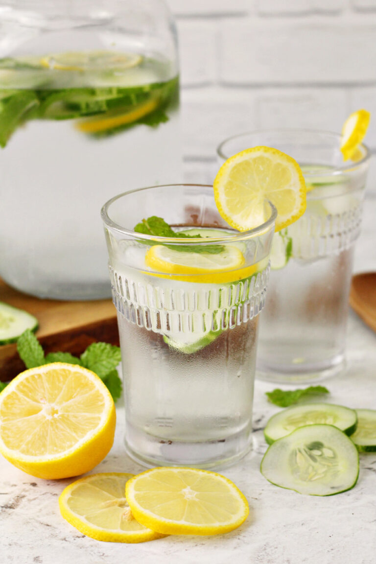Refreshing Cucumber, Lemon, and Mint Water served in glasses. Slices of lemons and cucumber sit in photo. Pitcher of infused water sits in background.