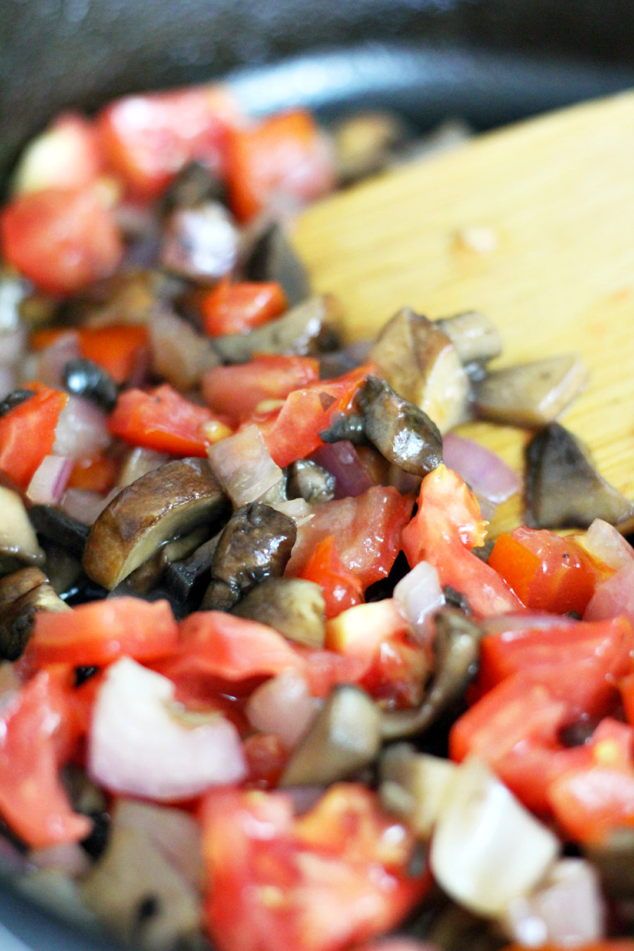 Chopped baby bella mushrooms, red onion, and tomato in a cast iron skillet.  Wooden spatula sits in skillet.