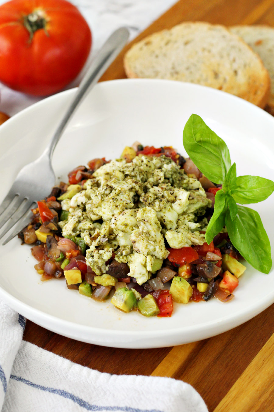 Pesto Infused Egg White Skillet served on white plate sits on top of wooden board. Silver fork sits on plate as well as fresh basil garnish. Tomato and rye toast are also in photo.