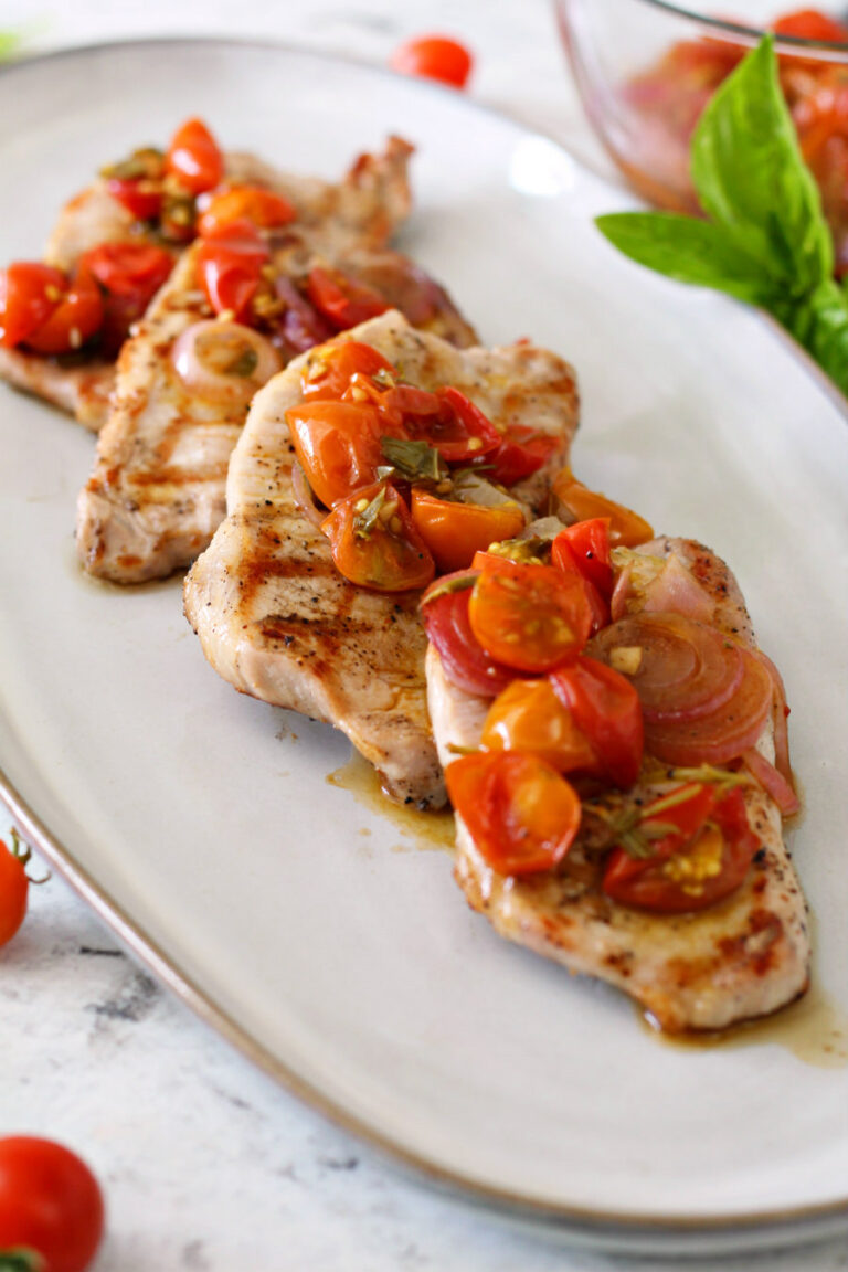 Grilled Boneless Pork Chops with Tomato Compote plated on a narrow gray oval plate. Bowl of tomato compote and fresh herbs also sit in photo.