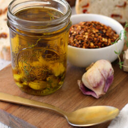 Mason jar of Spicy Garlic Confit Oil sits on wooden board with gold spoon, heads of garlic, fresh thyme, small bowl of red pepper flakes, and rustic bread in background.