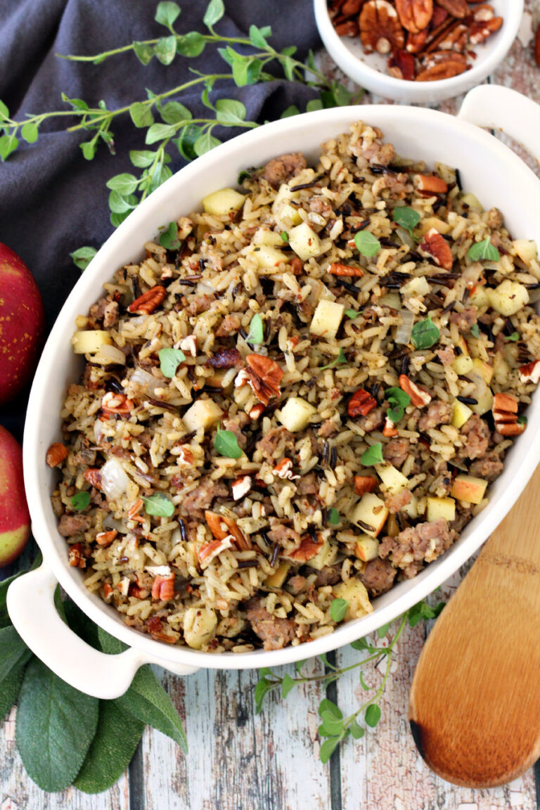 Flat lay of Sausage and Apple Wild Rice in white baking dish. Wooden spoon, small white bowl of pecans, fresh oregano, fresh sage, apples, and gray kitchen towel also in view.