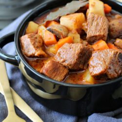 Partial view of Traditional Samhain Beef Stew in shallow black bowl. Bowl sits on gray kitchen towel with gold utensils, biscuits, and black lid with skull on top in background.