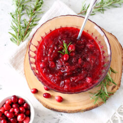 Top view of Rosemary and Bourbon Cranberry Sauce in glass serving bowl. Sauce is garnished with tiny sprig of rosemary. Silver spoon sits inside bowl. Bowl is on wood slice and white kitchen towel. Fresh rosemary sprigs and small bowl of fresh cranberries also in view.