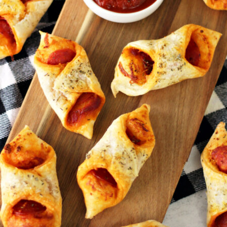 Flat lay view of Pepperoni Pizza Puff Pastry Bundles on wooden board. Black and white check kitchen towel and small white bowl of pizza sauce also in view.