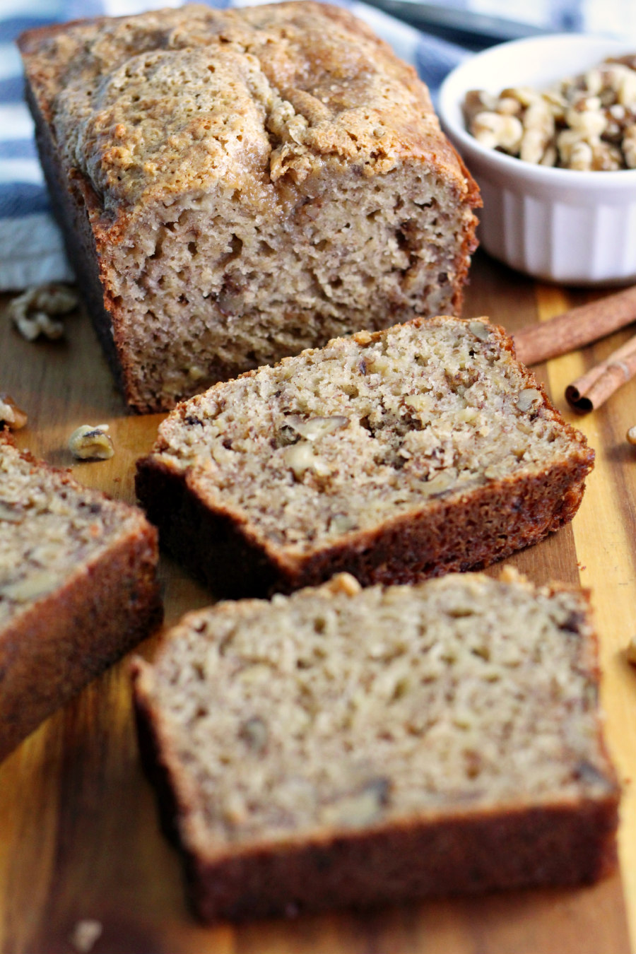 Greek Yogurt Banana Nut Bread loaf sits in back with three slices in front. Bread sits on wooden board along with walnuts, bowl of walnuts, cinnamon sticks, and blue and white striped kitchen towel in partial view in background.