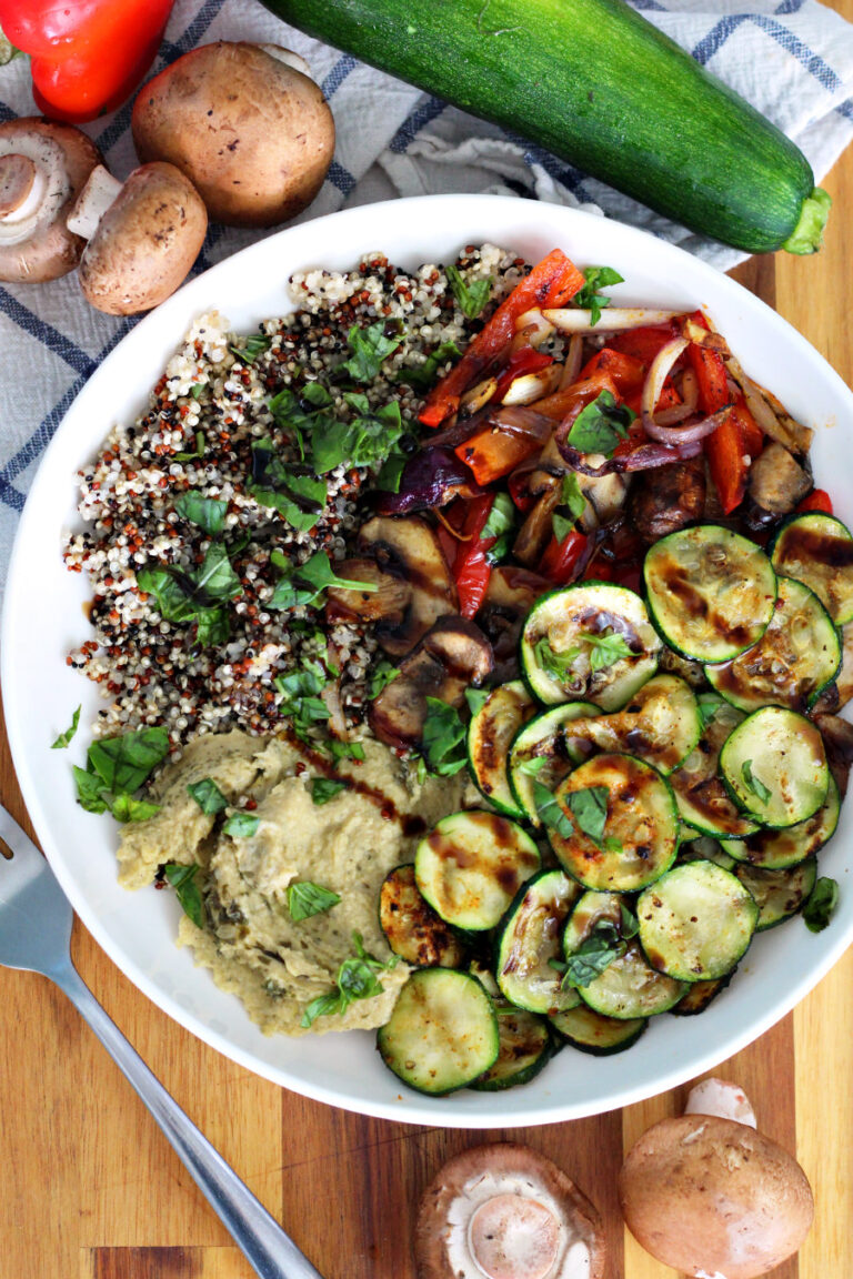 Top view of Grilled Vegetable Buddha Bowl in white serving dish. Dish sits on top of wooden board. Fork, baby bella mushrooms, blue and white kitchen towel, top of red bell pepper, and zucchini in partial view.
