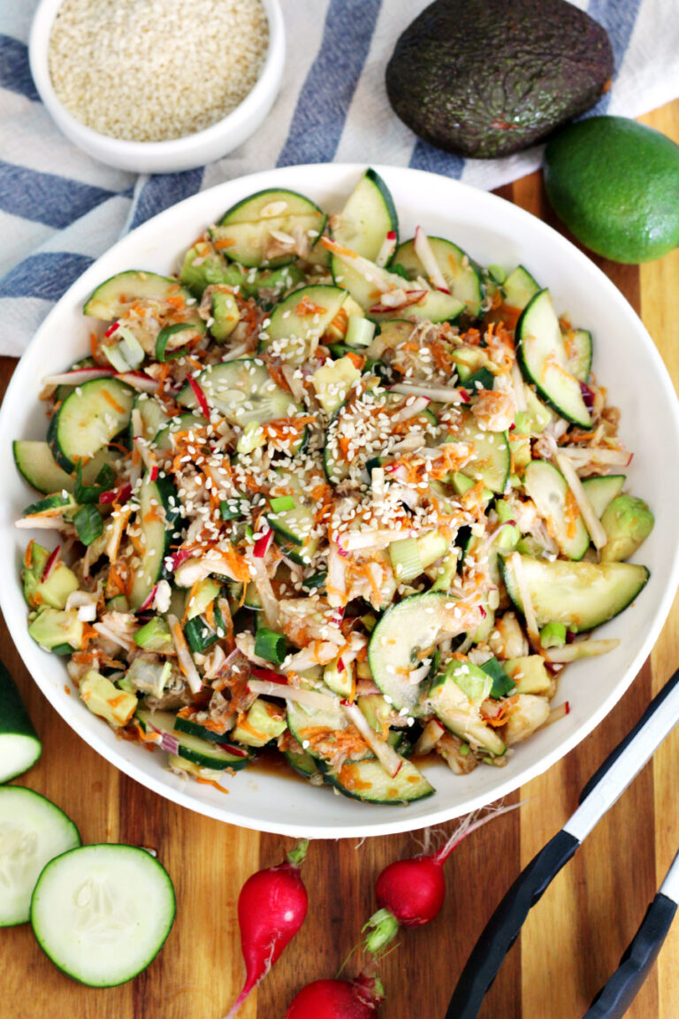 Top view of Crunchy Asian-Inspired Cucumber and Crab Salad in white serving dish. Dish sits on wooden board along with cucumber slices, radishes, metal tongs, a lime, an avocado, a small bowl of sesame seeds, and a striped kitchen towel.