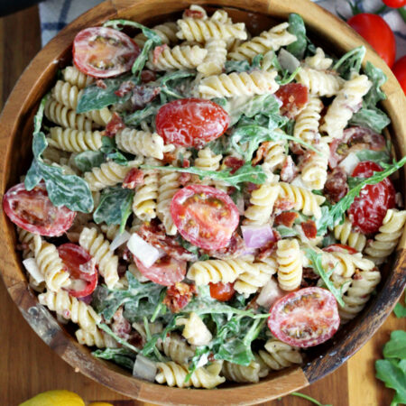 Top view of BLT Pasta Salad in wooden serving bowl on top of wooden board. Lemon, arugula, cherry tomatoes, tongs, and kitchen towel also in partial view.